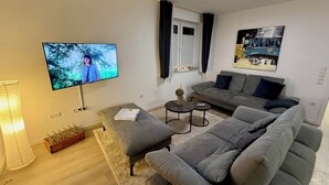 En stue med en grå sofa, et fladskærms-tv, et lille bord og en pyntende plante.