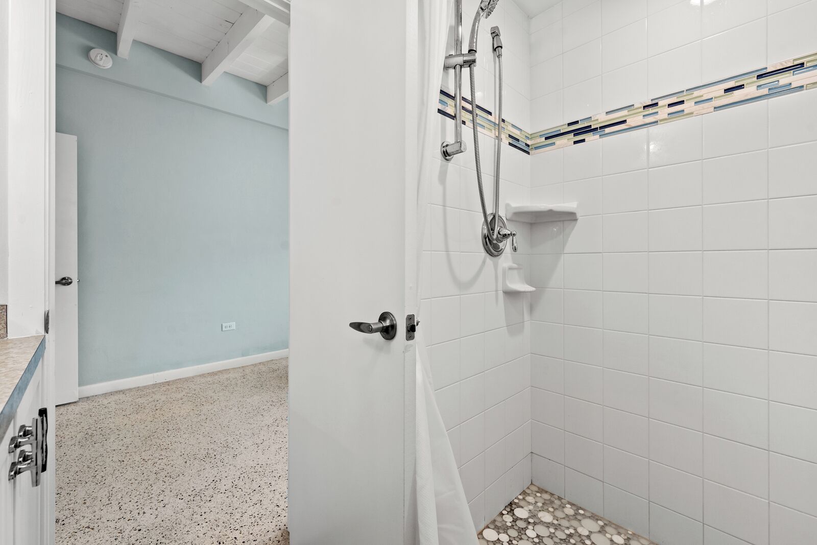 A bathroom with white tiled walls, a shower with a handheld showerhead, and a door with a modern handle.