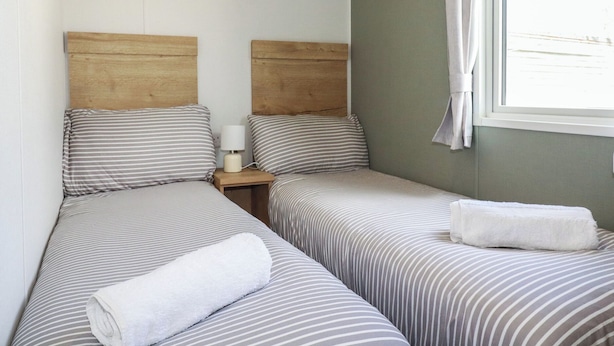 A room with two single beds, a nightstand with a lamp, and a window with a curtain. - Shamrocks Cove Caravan (Newquay)