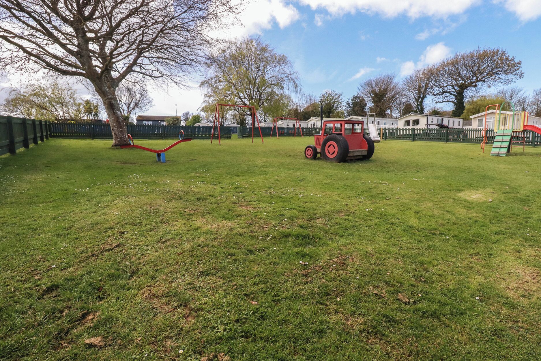 A playground with swings, a red tractor, and a fenced area.