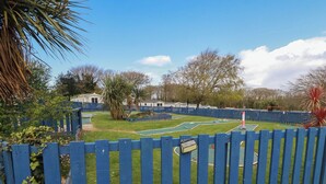 A fenced outdoor area with a green lawn, a small pool, and a tennis court. - Shamrocks Cove Caravan (Newquay)