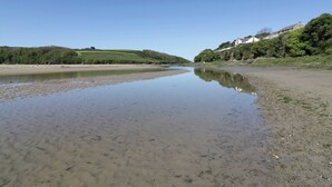 A river with calm water reflecting the sky, surrounded by green hills and a building on the right. - Shamrocks Cove Caravan (Newquay)