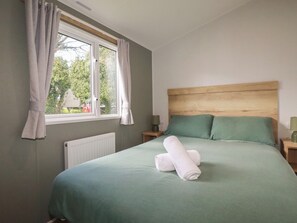 A bedroom with a bed, green bedding, a wooden headboard, a window with curtains, and a radiator. - Shamrocks Cove Caravan (Newquay)