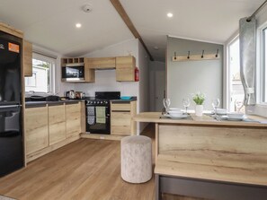 A modern kitchen with wooden cabinets, a built-in oven, and a dining area with a table set for two. - Shamrocks Cove Caravan (Newquay)