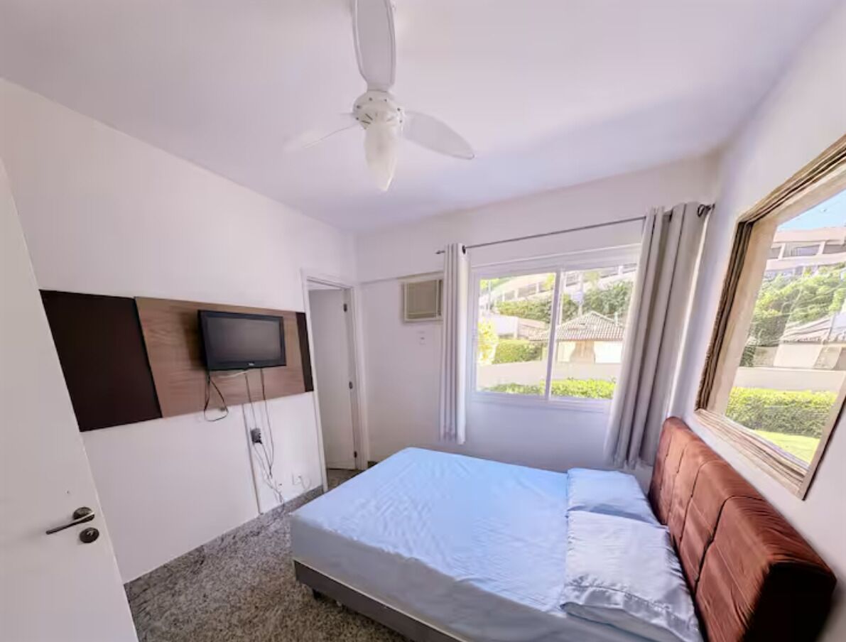 Une chambre à coucher avec un lit, un ventilateur de plafond, un téléviseur fixé au mur et une fenêtre avec des rideaux.
