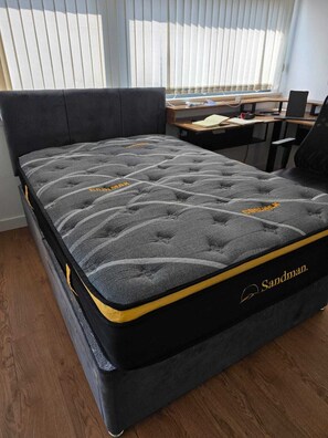 A bed with a mattress labeled 'Sandman,' a wooden desk, and a window with blinds. - Charming 2 bedroom apartment in Glasgow. Right next to the SEC and OVO Hydro. (Glasgow)