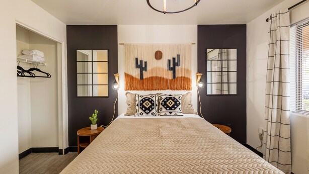 Ein Schlafzimmer mit einem großen Bett, einem Nachttisch mit Lampe, einem an der Wand befestigten Spiegel und einem Fenster mit Vorhängen.