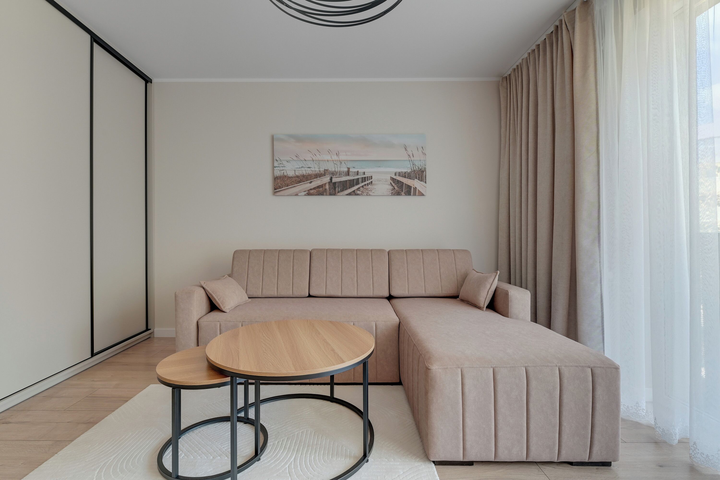 A modern living room with a beige sectional sofa, a round wooden coffee table, and a beach-themed painting on the wall.