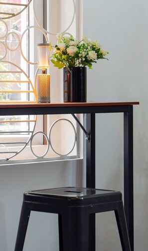 Un taburete negro, un jarrón negro con flores blancas y una lámpara metálica sobre una mesa de madera junto a una ventana con barandilla de metal.