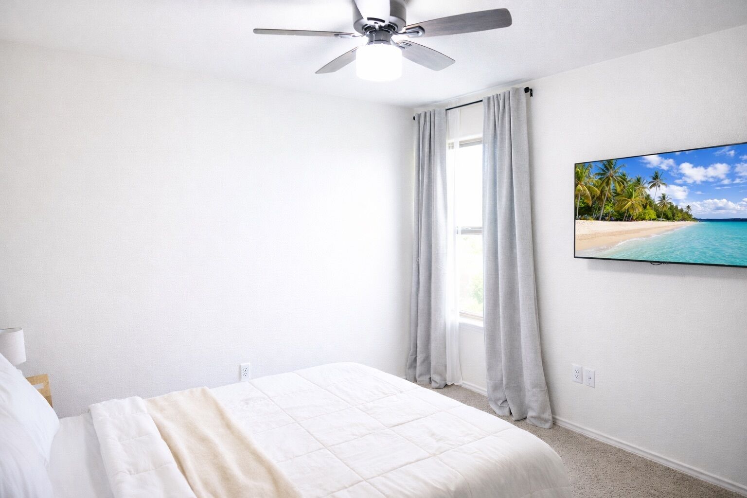 A bedroom with a bed, a ceiling fan, a window with curtains, and a wall-mounted TV displaying a beach scene.