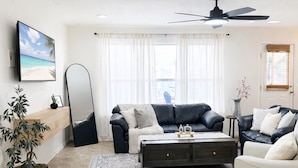 Un salon comprenant un canapé, une table basse, un téléviseur, un ventilateur de plafond et une fenêtre avec des rideaux.
