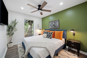 Une chambre à coucher avec un lit, un ventilateur de plafond, une table de chevet, une lampe, une plante en pot et une vue sur l’extérieur.