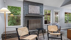 Un salon moderne avec une cheminée en pierre, deux fauteuils, une lampe sur pied et de grandes fenêtres donnant sur un paysage verdoyant.