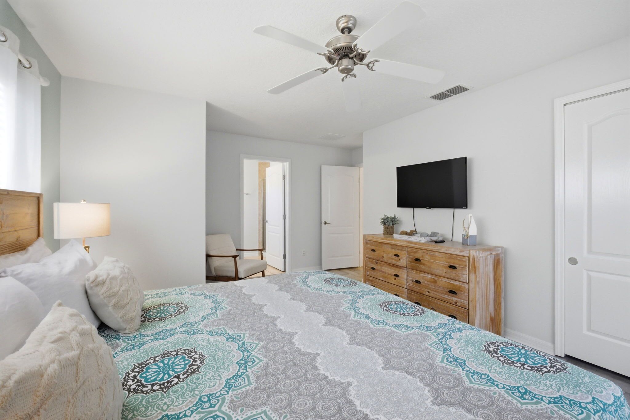 A bedroom with a bed, a dresser, a ceiling fan, a TV, and a door leading to another room.