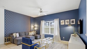 Ein Wohnzimmer mit einer Sofagarnitur, einem Couchtisch, einem blauen Sessel, einem Deckenventilator und einem Fenster mit Vorhängen.