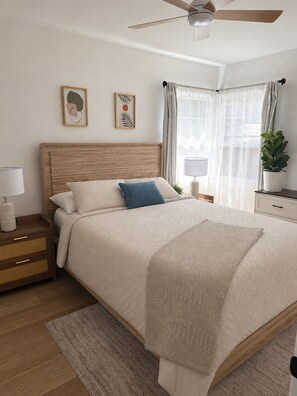 Un dormitorio con cabecera de madera, cama con manta color beige, mesita de noche con lámpara, mesita auxiliar, ventilador de techo y ventana con cortinas traslúcidas.