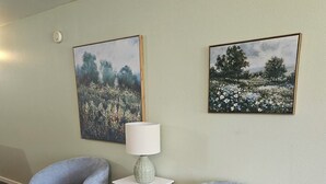 A room with two grey armchairs, a white side table with a lamp, and two landscape paintings on the wall.