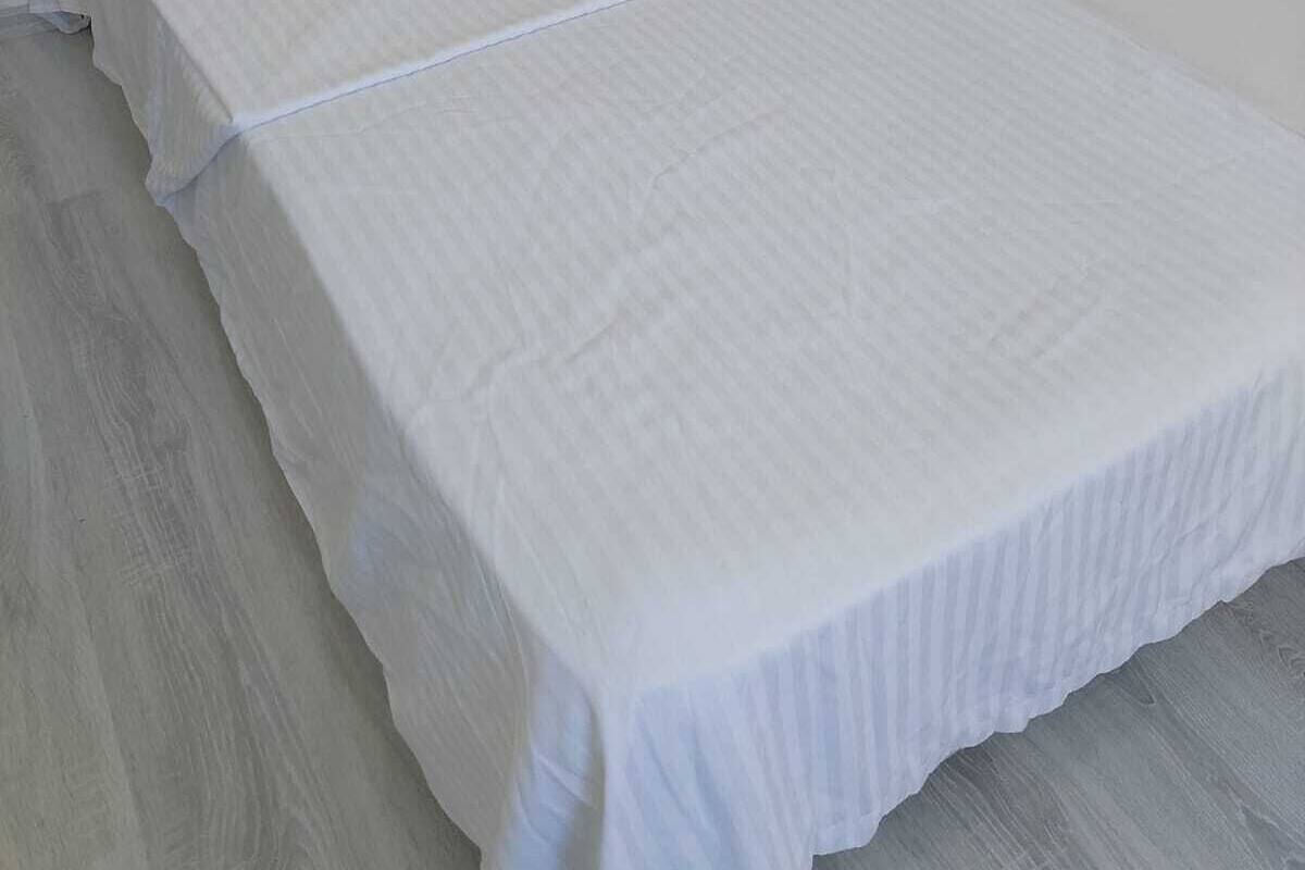A single bed with white bedding in a room with a plain white wall and a light-colored floor.
