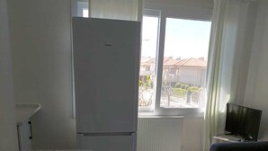 A room with a bed, a table with a lace tablecloth, a refrigerator, a television, and a window with curtains.