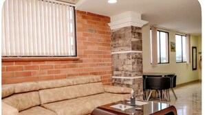 Un salon comprenant un canapé d’angle, une table basse avec un plateau en verre, un mur décoratif en briques et un coin repas avec une table et des chaises.