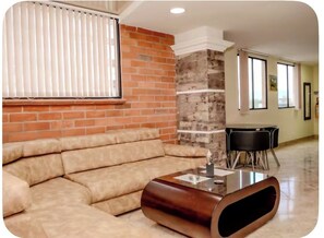 A living room with a sectional sofa, a glass-top coffee table, a brick accent wall, and a dining area with a table and chairs. - 108 LOVELY 2 BEDROOM APARTMENT NEAR PARK POBLADO (Medellín)