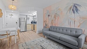 Een moderne woonkamer met een grijze bank, een eettafel met stoelen, een keuken met een koelkast en magnetron, en een wanddecoratie met een tropisch thema.