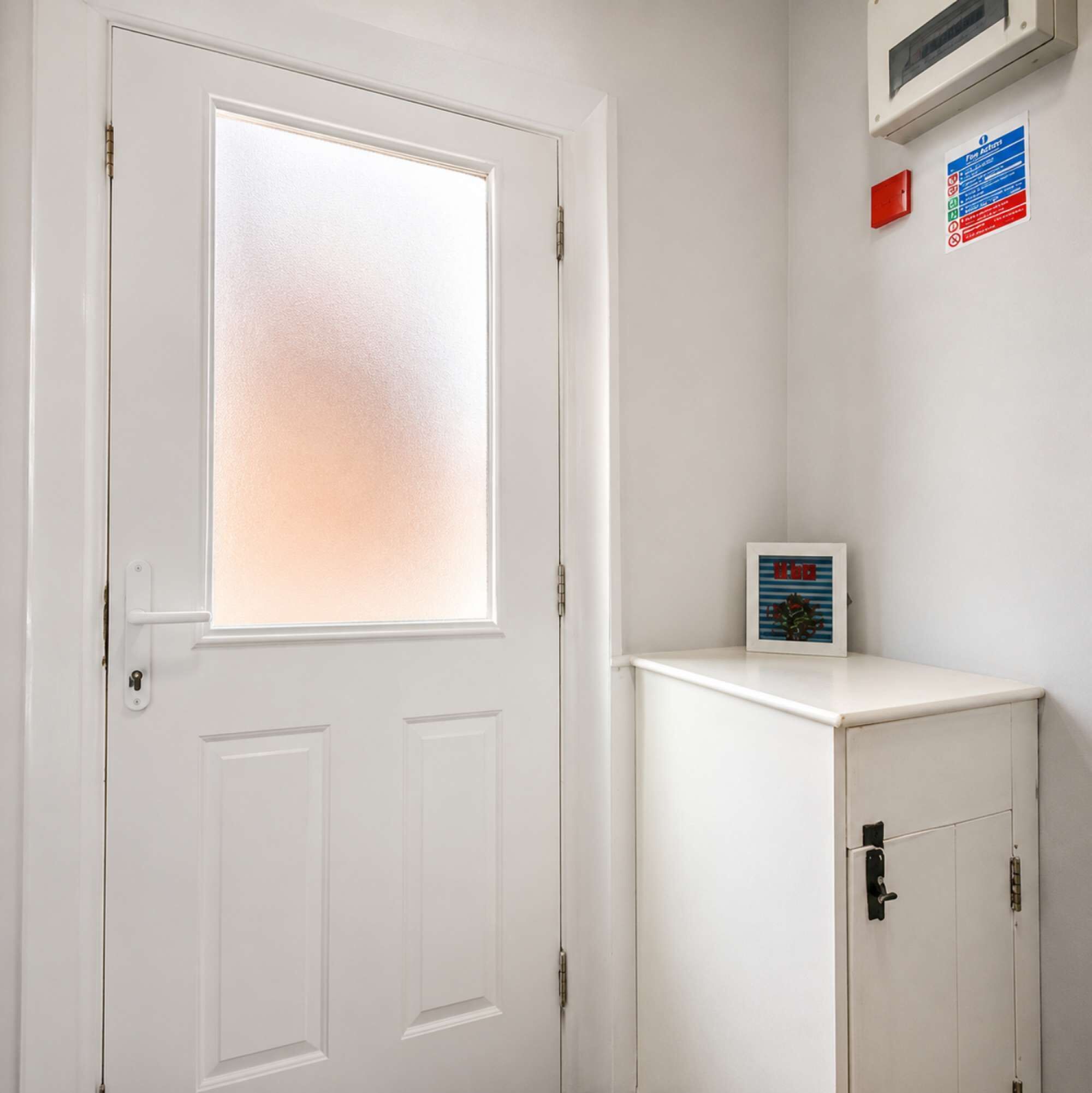 A white door with a frosted glass panel, a small cabinet with a framed picture, and a wall-mounted air conditioning unit.