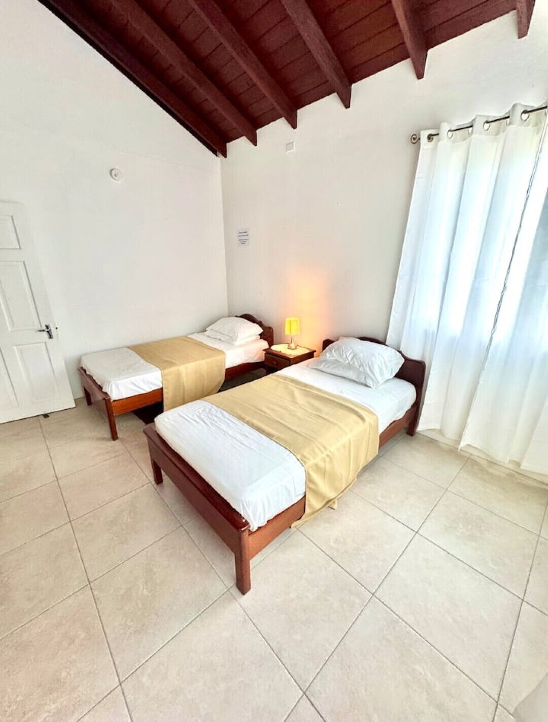 A room with two wooden beds, a nightstand with a lamp, white curtains, and a tiled floor.