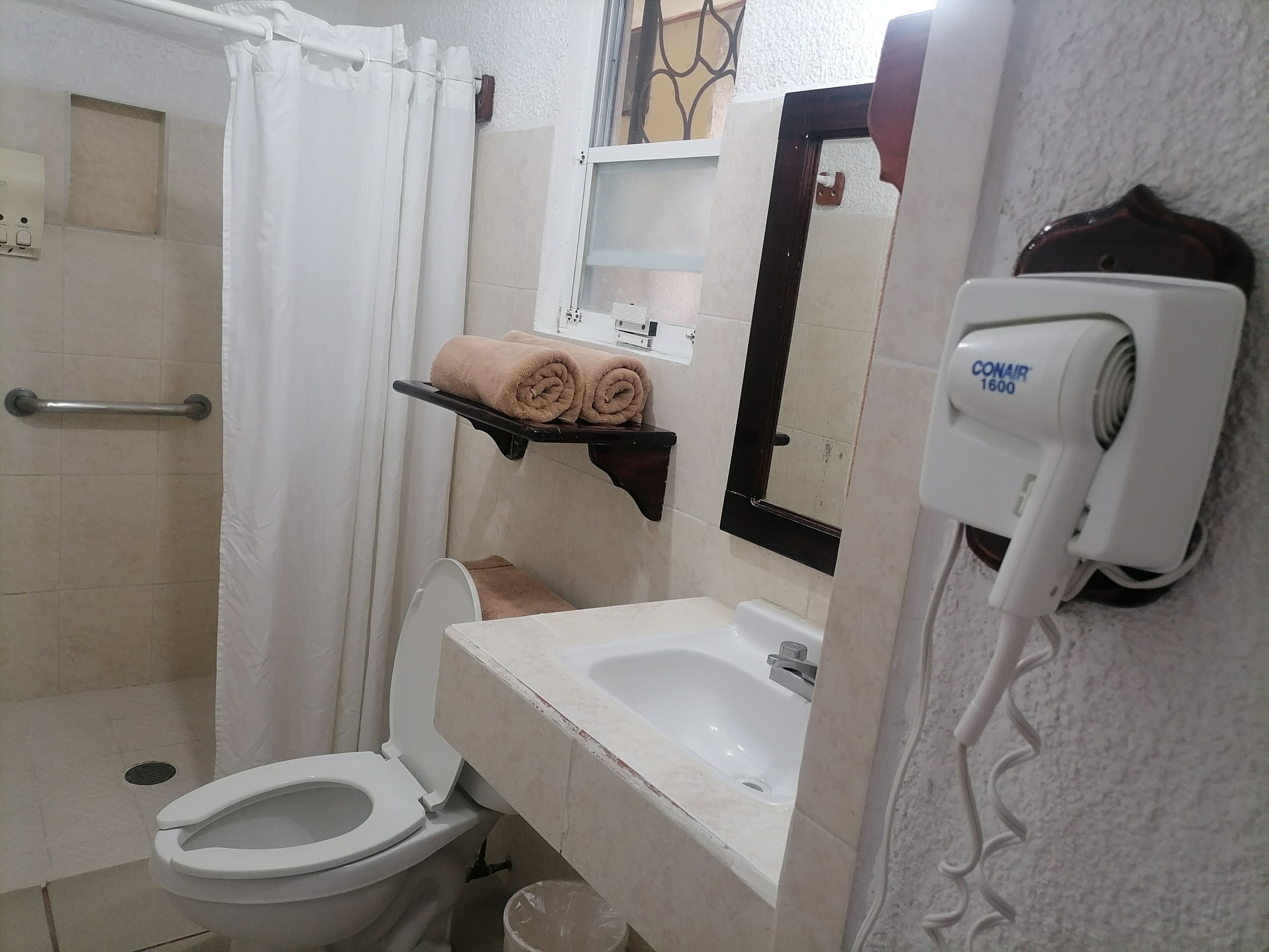 Un bany amb vàter, rentamig, dutxa i un assecador de cabells penjat a la paret.