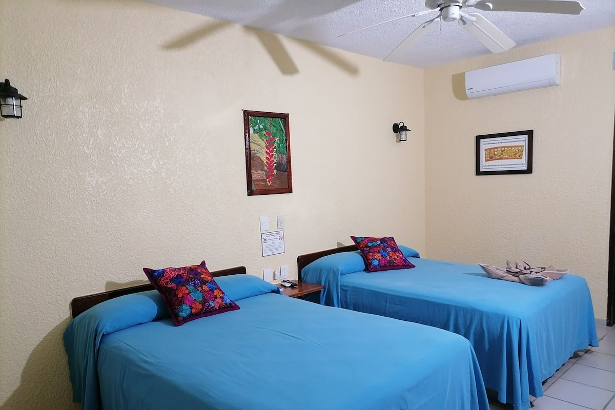 Habitación de hotel con dos camas, cada una con cubrecama azul y almohada colorida. Se observa un ventilador de techo, un equipo de aire acondicionado y dos lámparas fijadas a la pared.