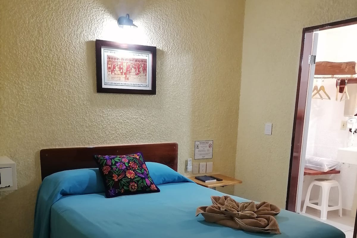 Una habitación de hotel con una cama, un cuadro en la pared, una mesita de noche y un baño visible a través de una puerta abierta.