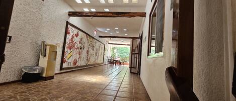 Un pasillo con baldosas estampadas, un cubo de basura sujeto a la pared, una puerta con un número y una ventana.