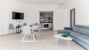 Een moderne woonkamer met een flatscreen-tv, een eethoek met stoelen en een tafel, een grijze bank en een keuken die door een open doorgang zichtbaar is.