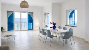 Een moderne eetruimte met een witte tafel, grijze stoelen, een blauwe vaas met bloemen en grote ramen met blauwe luiken.