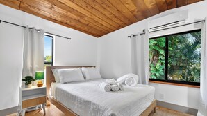 Ein Schlafzimmer mit Holzboden, einem Bett mit weißer Bettwäsche, einem Nachttisch mit Pflanze und einem Fenster mit Blick ins Grüne.