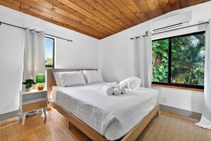 Ein Schlafzimmer mit Holzboden, einem Bett mit weißer Bettwäsche, einem Nachttisch mit Pflanze und einem Fenster mit Blick ins Grüne.