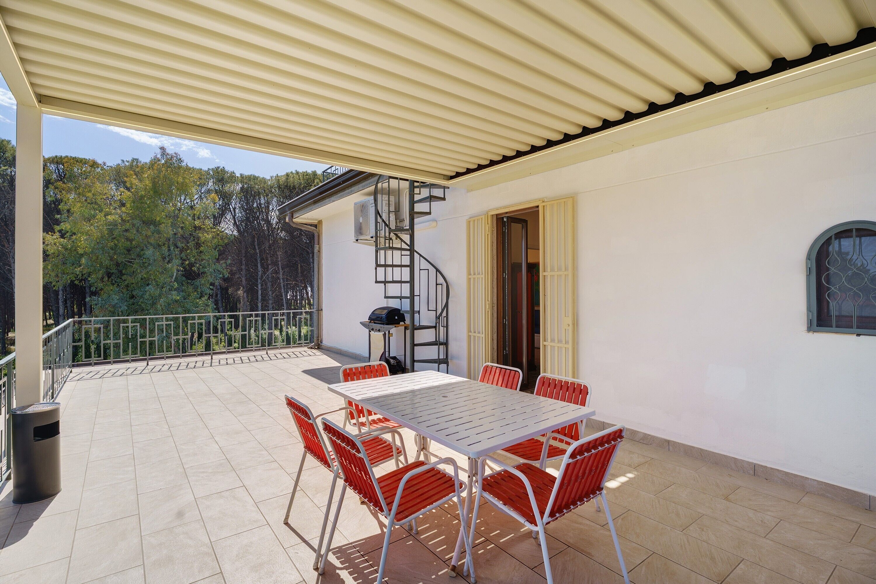 A covered patio with a table and chairs, a spiral staircase, and a view of trees.