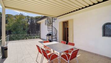 A covered patio with a table and chairs, a spiral staircase, and a view of trees.