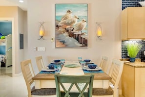 A dining area with a table set for four, two wall-mounted light fixtures, and a painting of seagulls on the wall. - Beachside Condo w/ Pools, Hot Tubs & Balcony (South Padre Island)