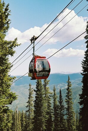 Un teleférico con la leyenda 'ASPEN' escrito en él, suspendido por cables, rodeado de árboles y montañas.