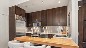 Une cuisine moderne avec des meubles en bois foncé, des appareils en acier inoxydable, un îlot central en bois doté de tabourets, et un micro-ondes encastré.