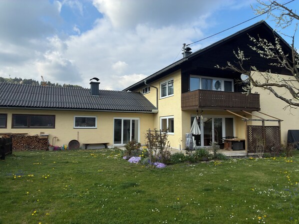 A two-story house with a balcony, a garden with flowers, and a clear sky.