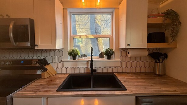 Una cocina con un lavamanos negro, encimera de madera, armarios blancos, una ventana con plantas y un colgante.