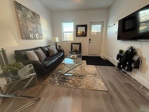 A living room with a black leather sofa, a glass coffee table, a TV mounted on the wall, and a door with a window. - 3BDR Entire House 1BLK to River/5min Walk to DTWN (Saskatoon)