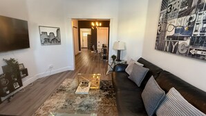 Una sala de estar con un sofá de cuero negro, una mesa de centro de vidrio, un cuadro con temática urbana y un televisor de pantalla plana montado en la pared.