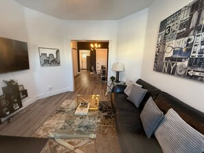A living room with a black leather sofa, a glass coffee table, a city-themed wall art, and a flat-screen TV mounted on the wall. - 3BDR Entire House 1BLK to River/5min Walk to DTWN (Saskatoon)