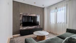 Una sala de estar moderna con un televisor de pantalla plana montado en una pared con textura similar al mármol, un mueble de entretenimiento oscuro, una mesa de centro redonda con base de madera y un sofá gris.