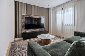 Ein modernes Wohnzimmer mit einem an der Wand mit Marmoroptik befestigten Flachbildfernseher, einer dunklen Unterhaltungsecke, einem runden Couchtisch mit Holzfuß und einem grauen Sofa.