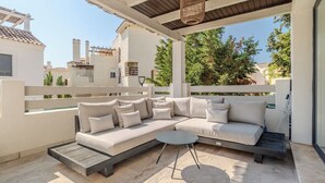 Un espacio de estar al aire libre techado con un sofá y una mesita, un pérgola arriba y vistas a edificios y árboles.