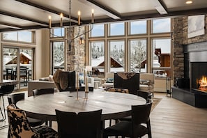 A spacious living room with a large round table, stone fireplace, and large windows offering a view of snow-covered cabins. - *New Build* Black Eagle Ski-In/Ski-Out Retreat (Big Sky)